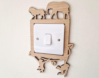 Può includere: Una placca per interruttore della luce in legno chiaro con sagome di animali tagliate al laser, tra cui un leone e un elefante in alto e scimmie appese a liane in basso, che circondano un interruttore bianco standard con due viti metalliche.