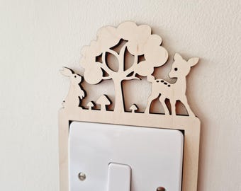 May include: A light-colored wooden light switch cover plate featuring a laser-cut design of a tree, a standing rabbit, a fawn, and small mushrooms. The decorative wooden cutout is mounted around a standard white rectangular light switch.