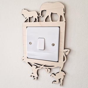 May include: A wooden light switch cover with animal cutouts. The design includes a lion, elephant, and monkeys. The cover is made of light-colored wood and surrounds a white light switch. The overall shape is rectangular.