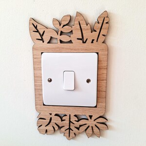 May include: A decorative wooden light switch cover featuring a leaf design. The light switch cover is made of light brown wood and surrounds a white light switch. The leaf design is cut out of the wood.