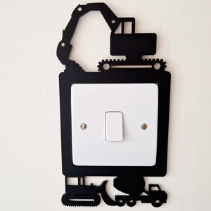 May include: Black metal light switch cover with a construction vehicle theme. The cover features a silhouette of an excavator, a bulldozer, and a dump truck. The white light switch is centered within the black frame.