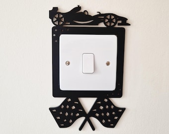 May include: A black decorative light switch cover plate featuring a silhouette of a race car at the top and two crossed checkered flags at the bottom. The square frame includes small star cutouts and surrounds a standard white rectangular light switch.