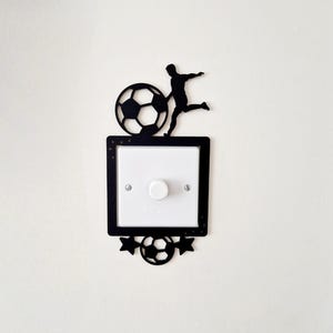 May include: A white light switch with a circular knob, framed by a black decorative cover. The cover features a silhouette of a footballer kicking a ball at the top, and a smaller football flanked by two stars at the bottom edge.