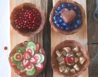 May include: Four mini pies with different fillings. The first pie has a red berry filling, the second has a blueberry filling with a heart-shaped chocolate piece on top, the third has a strawberry and kiwi filling, and the fourth has a brown apple filling with gold flakes on top.