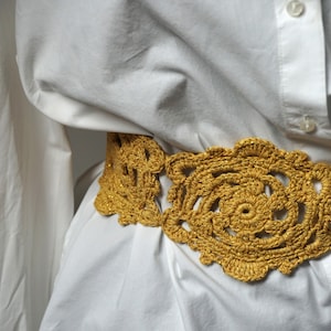 Puede incluir: Un cinturón de ganchillo dorado con un diseño floral. El cinturón se lleva sobre una camisa blanca abotonada. El intrincado patrón y el color cálido del cinturón crean un contraste llamativo con la tela blanca.
