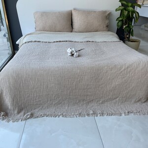 K&ouml;nnte beinhalten: Eine beige Waffel-Decke mit Fransen. Die Decke liegt auf einem Bett mit zwei beigen Kissen und einem kleinen Strau&szlig; wei&szlig;er Baumwolle.