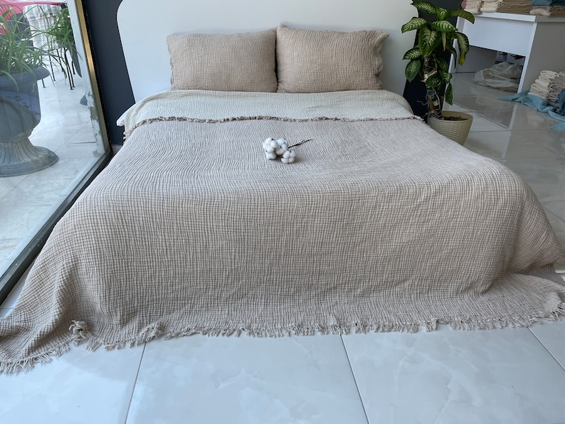 K&ouml;nnte beinhalten: Eine beige Waffel-Decke mit Fransen. Die Decke liegt auf einem Bett mit zwei beigen Kissen und einem kleinen Strau&szlig; wei&szlig;er Baumwolle.
