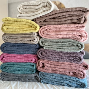 K&ouml;nnte beinhalten: Ein Stapel von 10 gefalteten Baumwoll-Musselin-Decken in verschiedenen Farben, darunter Beige, Braun, Gelb, Grau, Blau, Rosa und Gr&uuml;n.