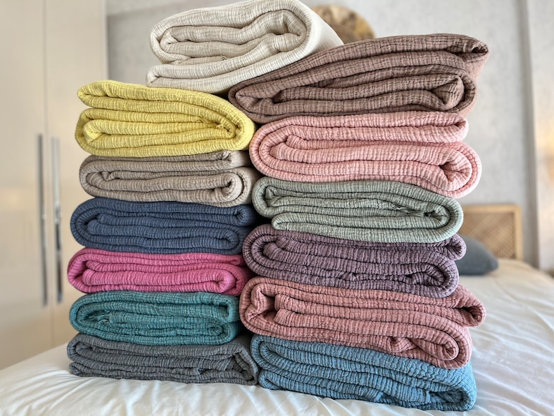 K&ouml;nnte beinhalten: Ein Stapel von 10 gefalteten Baumwoll-Musselin-Decken in verschiedenen Farben, darunter Beige, Braun, Gelb, Grau, Blau, Rosa und Gr&uuml;n.