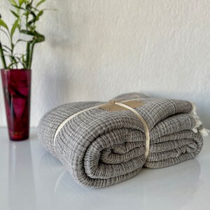 May include: A folded grey blanket with a textured weave, tied with a cream ribbon. The blanket has a fringed edge and a small tag. A tall, dark red vase with green bamboo stalks is in the background.