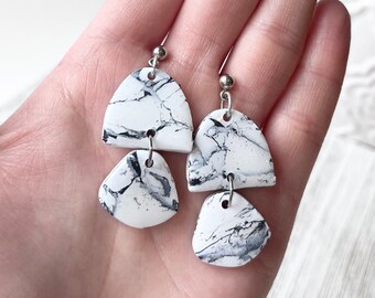 May include: A pair of handmade earrings featuring a marbled design in white and gray. Each earring has two geometric shapes, a half-circle and a triangle, connected by silver-colored hooks. The earrings are held in a hand against a soft background.
