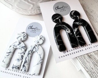 May include: A pair of black and white marble arch earrings. The earrings are made of polymer clay and have a black and white marble pattern. The earrings are about 2 inches long. The earrings are on a white card with the text "ChanRose Gems" and the social media handle "@chanrosegems".