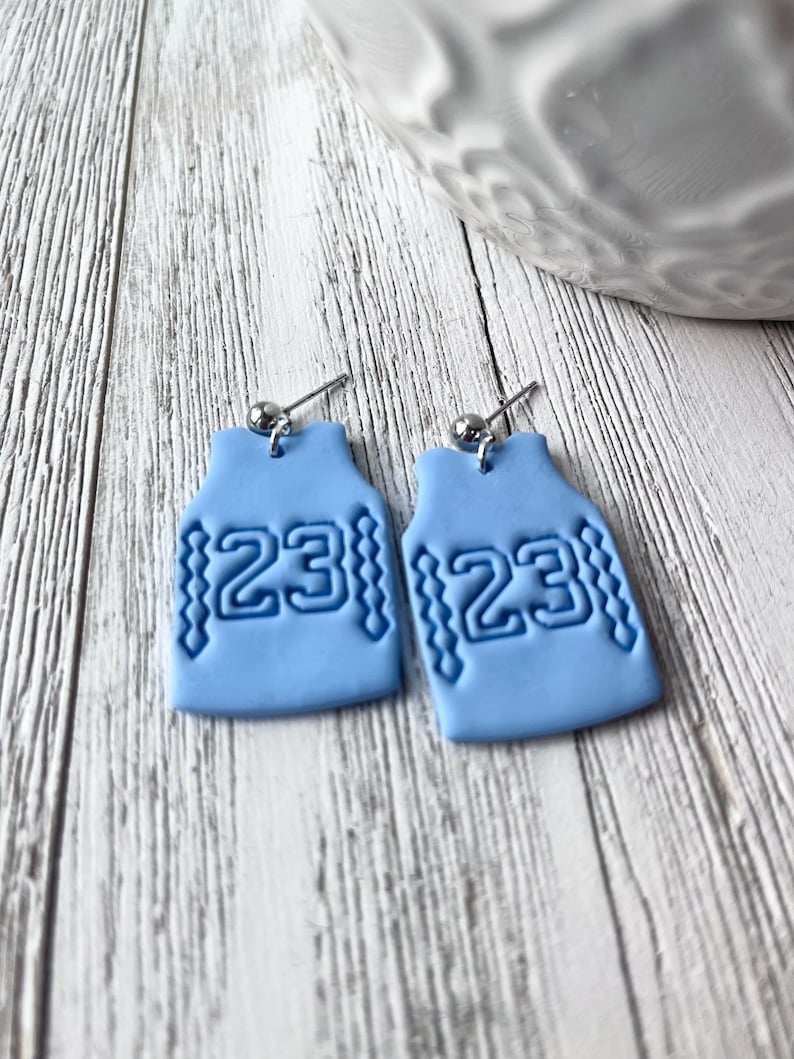 Op de afbeelding: Een paar lichtblauwe oorbellen in de vorm van basketbaltruitjes met het nummer 23 op elk. De oorbellen zijn gemaakt van polymer clay en hebben zilveren oorhaakjes.