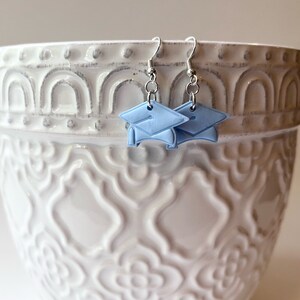 Peut inclure: Une paire de boucles d'oreilles en argent en forme de chapeau de graduation avec des capuchons bleu clair. Les boucles d'oreilles sont suspendues &agrave; un pot en c&eacute;ramique blanche avec un motif d&eacute;coratif.