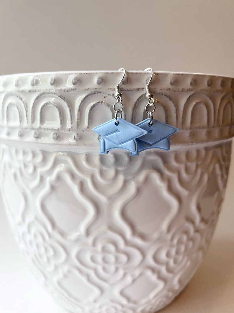Peut inclure: Une paire de boucles d'oreilles en argent en forme de chapeau de graduation avec des capuchons bleu clair. Les boucles d'oreilles sont suspendues &agrave; un pot en c&eacute;ramique blanche avec un motif d&eacute;coratif.
