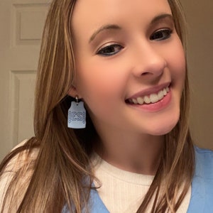 Op de afbeelding: Een vrouw draagt een lichtblauw scrubtop met een wit onderhemd. Ze draagt oorbellen in de vorm van een blauw vest met de letters "ESI" aan de voorkant.