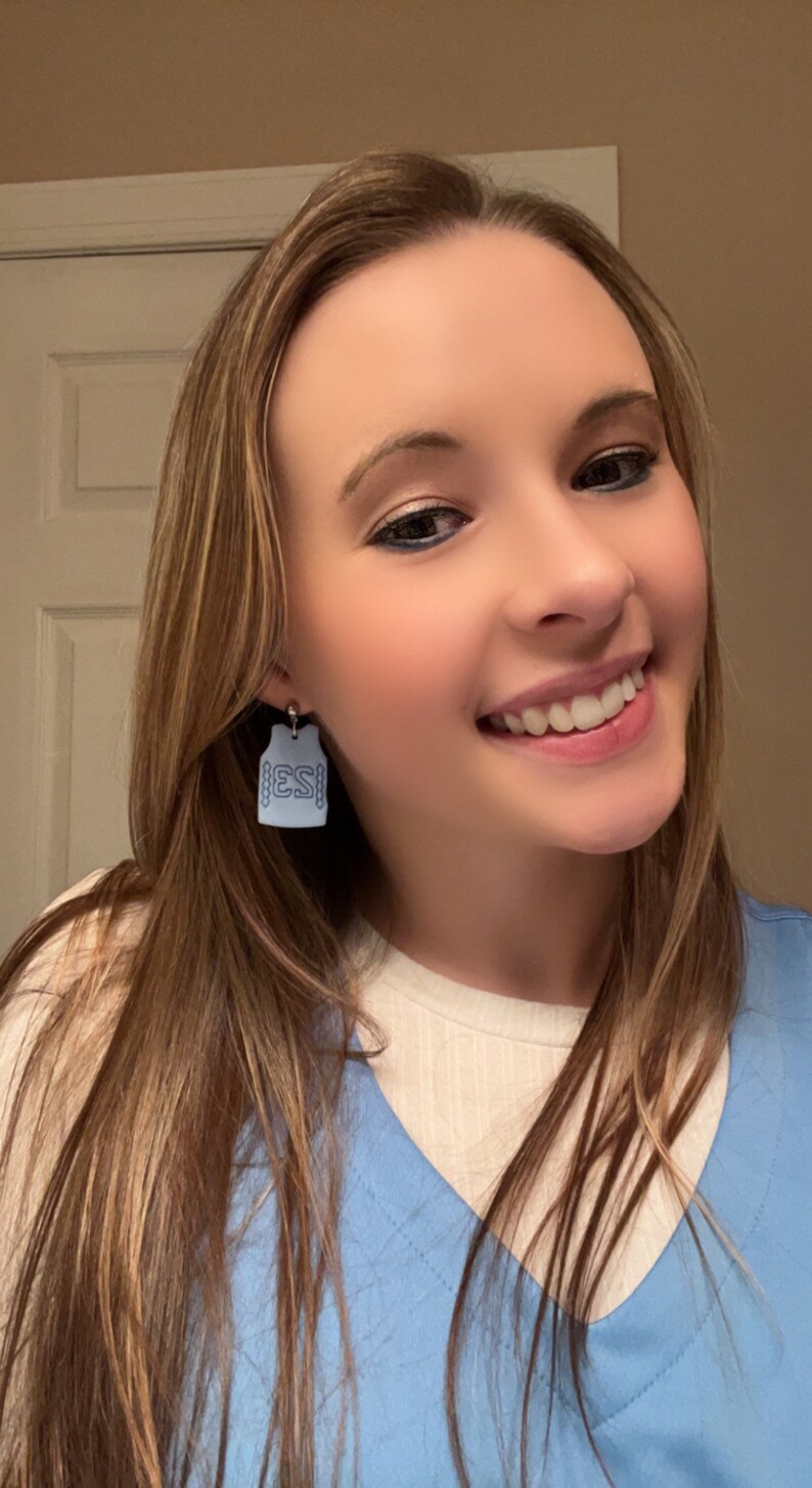 Op de afbeelding: Een vrouw draagt een lichtblauw scrubtop met een wit onderhemd. Ze draagt oorbellen in de vorm van een blauw vest met de letters "ESI" aan de voorkant.