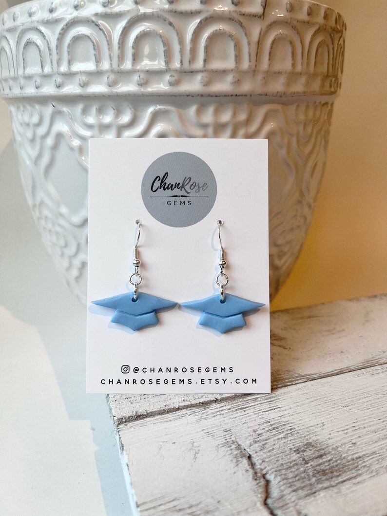 Peut inclure: Une paire de boucles d'oreilles en forme de chapeau de graduation bleu clair sur une carte blanche avec le texte "ChanRose Gems", le nom de profil sur les r&eacute;seaux sociaux "@chanrosegems" et le site web "chanrosegems.etsy.com".