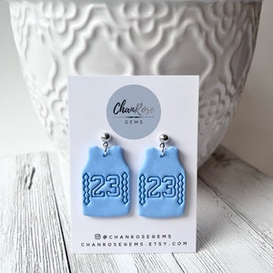 Op de afbeelding: Een paar lichtblauwe basketbaltrui-oorbellen met het nummer 23 op elk. De oorbellen zijn gemaakt van polymer clay en hebben zilveren haakjes.