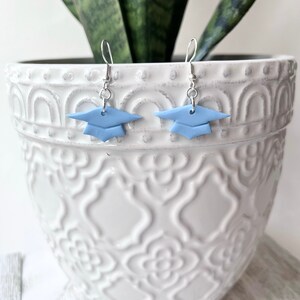 Peut inclure: Une paire de boucles d'oreilles en forme de chapeau de graduation bleu avec des crochets argent&eacute;s. Les boucles d'oreilles sont en p&acirc;te polym&egrave;re et constituent un moyen amusant et festif de c&eacute;l&eacute;brer la remise des dipl&ocirc;mes.