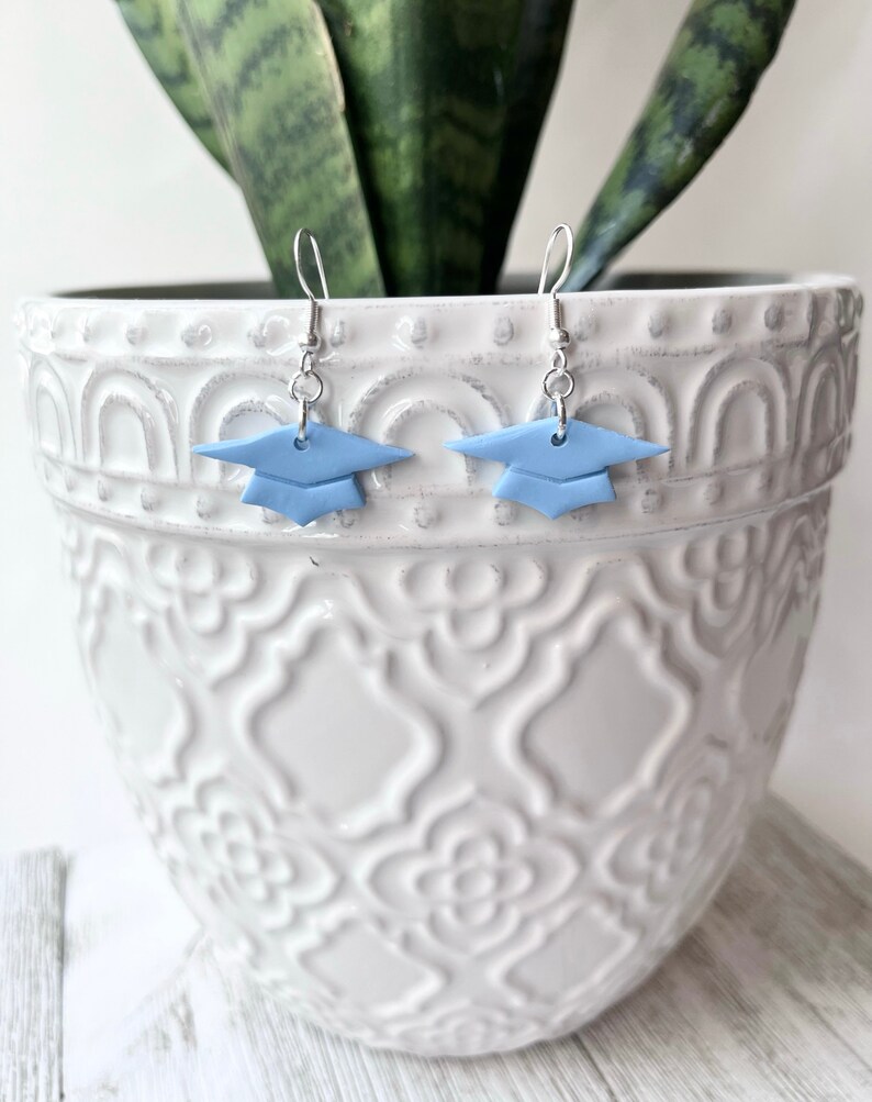 Peut inclure: Une paire de boucles d'oreilles en forme de chapeau de graduation bleu avec des crochets argent&eacute;s. Les boucles d'oreilles sont en p&acirc;te polym&egrave;re et constituent un moyen amusant et festif de c&eacute;l&eacute;brer la remise des dipl&ocirc;mes.