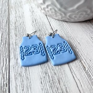 Op de afbeelding: Een paar lichtblauwe oorbellen in de vorm van basketbaltruitjes met het nummer 23 op elk. De oorbellen zijn gemaakt van polymer clay en hebben zilveren haakjes.