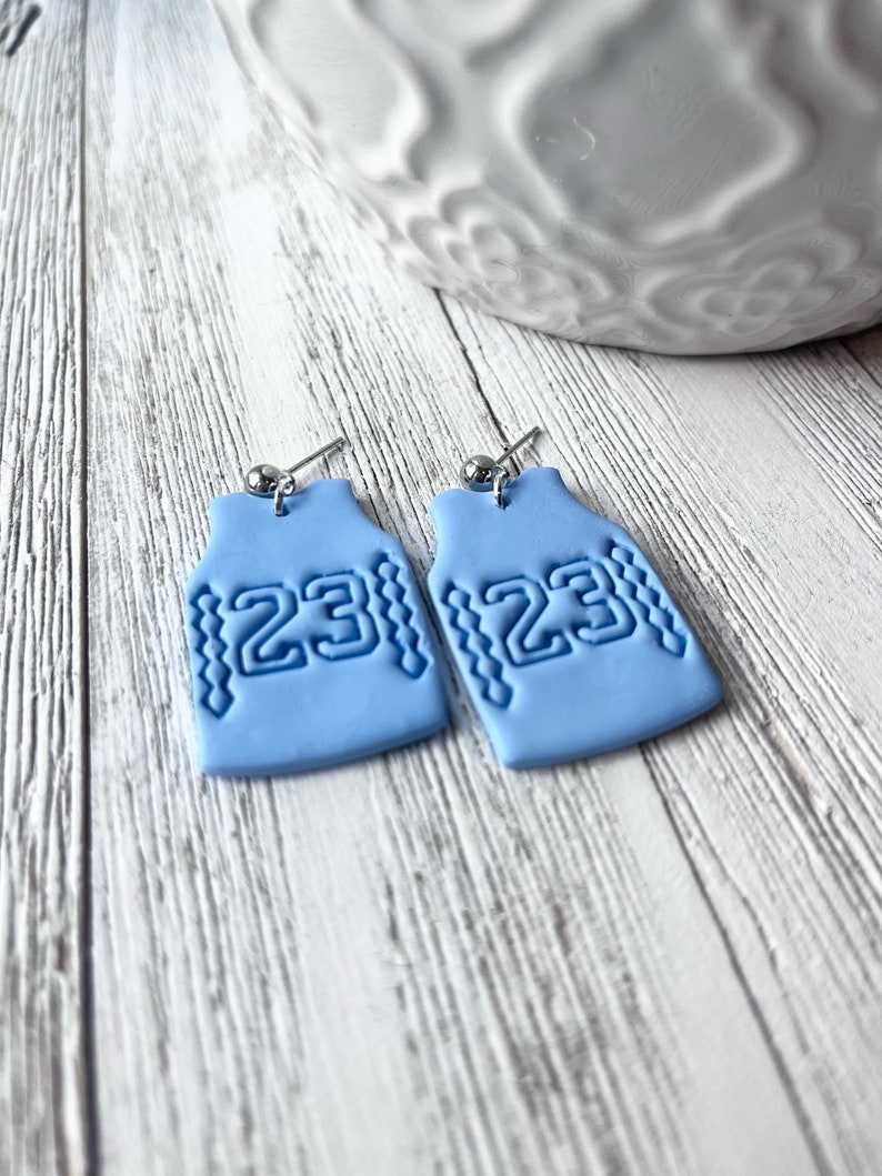 Op de afbeelding: Een paar lichtblauwe oorbellen in de vorm van basketbaltruitjes met het nummer 23 op elk. De oorbellen zijn gemaakt van polymer clay en hebben zilveren haakjes.