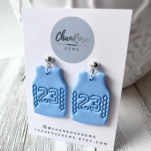 Op de afbeelding: Een paar lichtblauwe kleioorbellen in de vorm van basketbaltruitjes met het nummer 23 op elk. De oorbellen bevinden zich op een witte kaart met de tekst "ChanRose Gems" en de social media-handle "@chanrosegems".