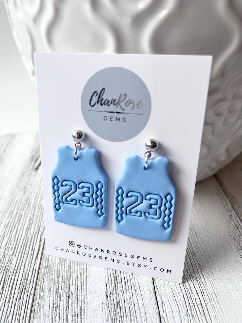 Op de afbeelding: Een paar lichtblauwe kleioorbellen in de vorm van basketbaltruitjes met het nummer 23 op elk. De oorbellen bevinden zich op een witte kaart met de tekst "ChanRose Gems" en de social media-handle "@chanrosegems".
