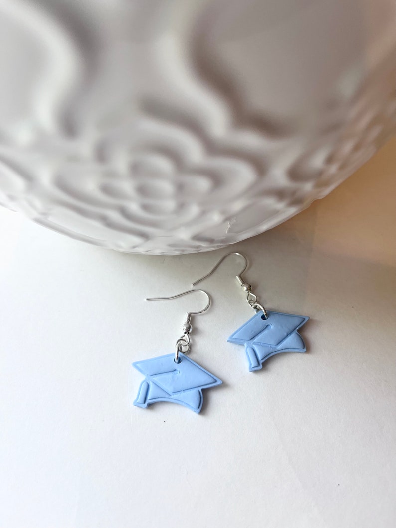 Peut inclure: Une paire de boucles d'oreilles pendantes argent&eacute;es avec des breloques de chapeau de graduation bleu clair. Les chapeaux sont en p&acirc;te polym&egrave;re et ont une finition mate.