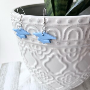 Peut inclure: Une paire de boucles d'oreilles en forme de chapeau de graduation bleu avec des crochets argent&eacute;s. Les boucles d'oreilles sont suspendues &agrave; un pot en c&eacute;ramique blanche avec un motif d&eacute;coratif.