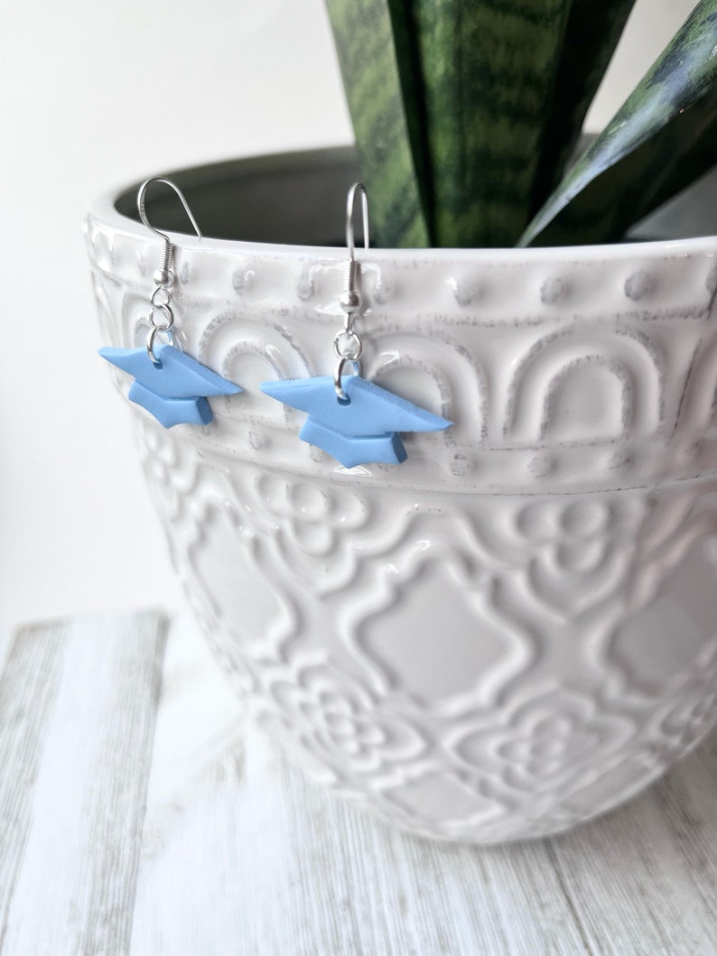 Peut inclure: Une paire de boucles d'oreilles en forme de chapeau de graduation bleu avec des crochets argent&eacute;s. Les boucles d'oreilles sont suspendues &agrave; un pot en c&eacute;ramique blanche avec un motif d&eacute;coratif.