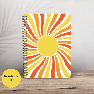 May include: A spiral-bound notebook with a sunburst design in yellow, orange, and white. The cover features a large yellow sun with radiating stripes. A yellow circle with the text "Notebook 5" is in the lower left corner.