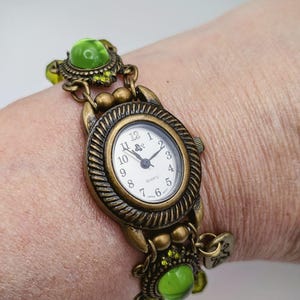 May include: An antique brass-toned quartz watch featuring an oval face with a white dial, black numerals, and hands. The watch band is composed of decorative metal links interspersed with round, translucent lime green cabochon beads.