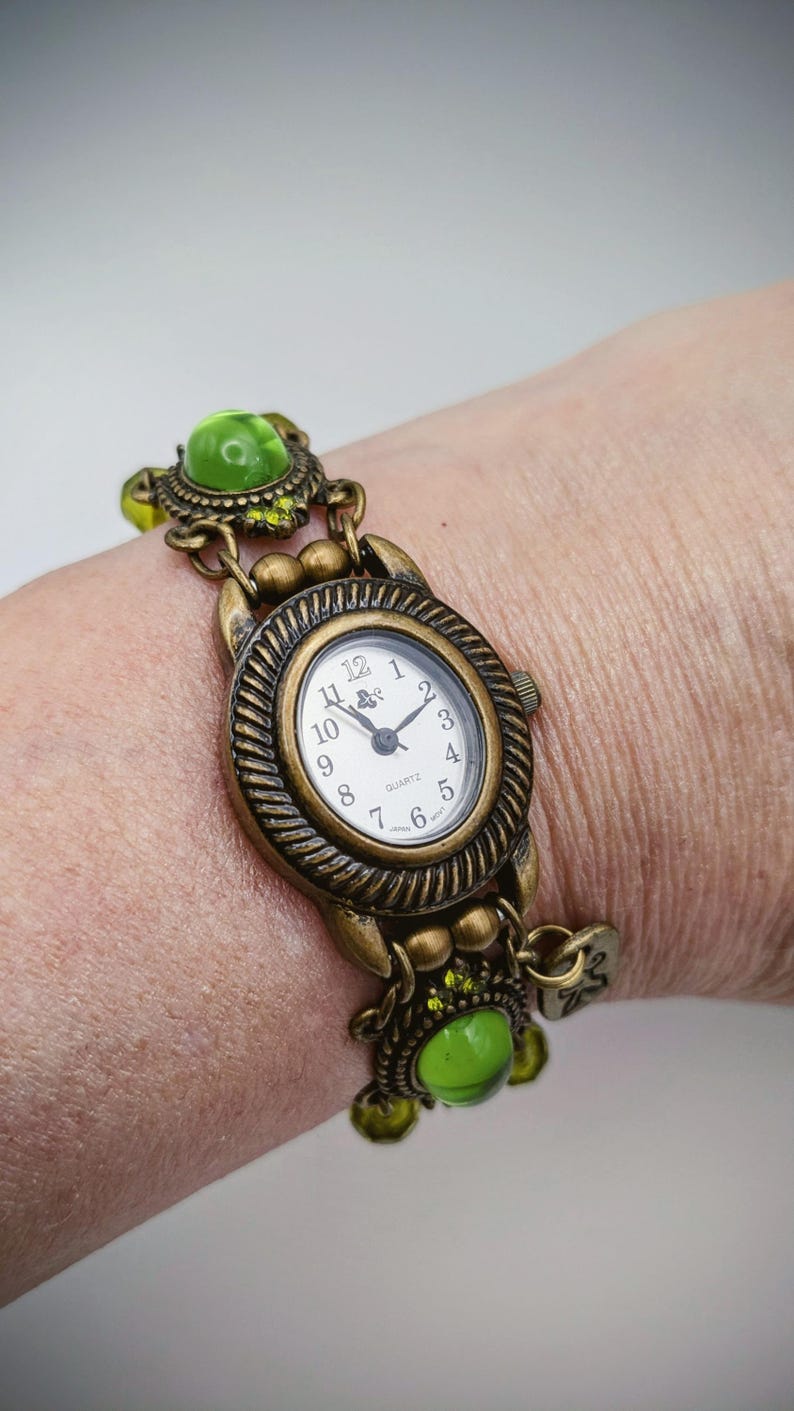 May include: An antique brass-toned quartz watch featuring an oval face with a white dial, black numerals, and hands. The watch band is composed of decorative metal links interspersed with round, translucent lime green cabochon beads.