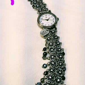 May include: A silver-toned quartz watch with a white face and black numerals. The band is composed of multiple chains adorned with small metal flower charms, clear crystals, and dangling black beads. A small rectangular metal tag hangs near the watch face.