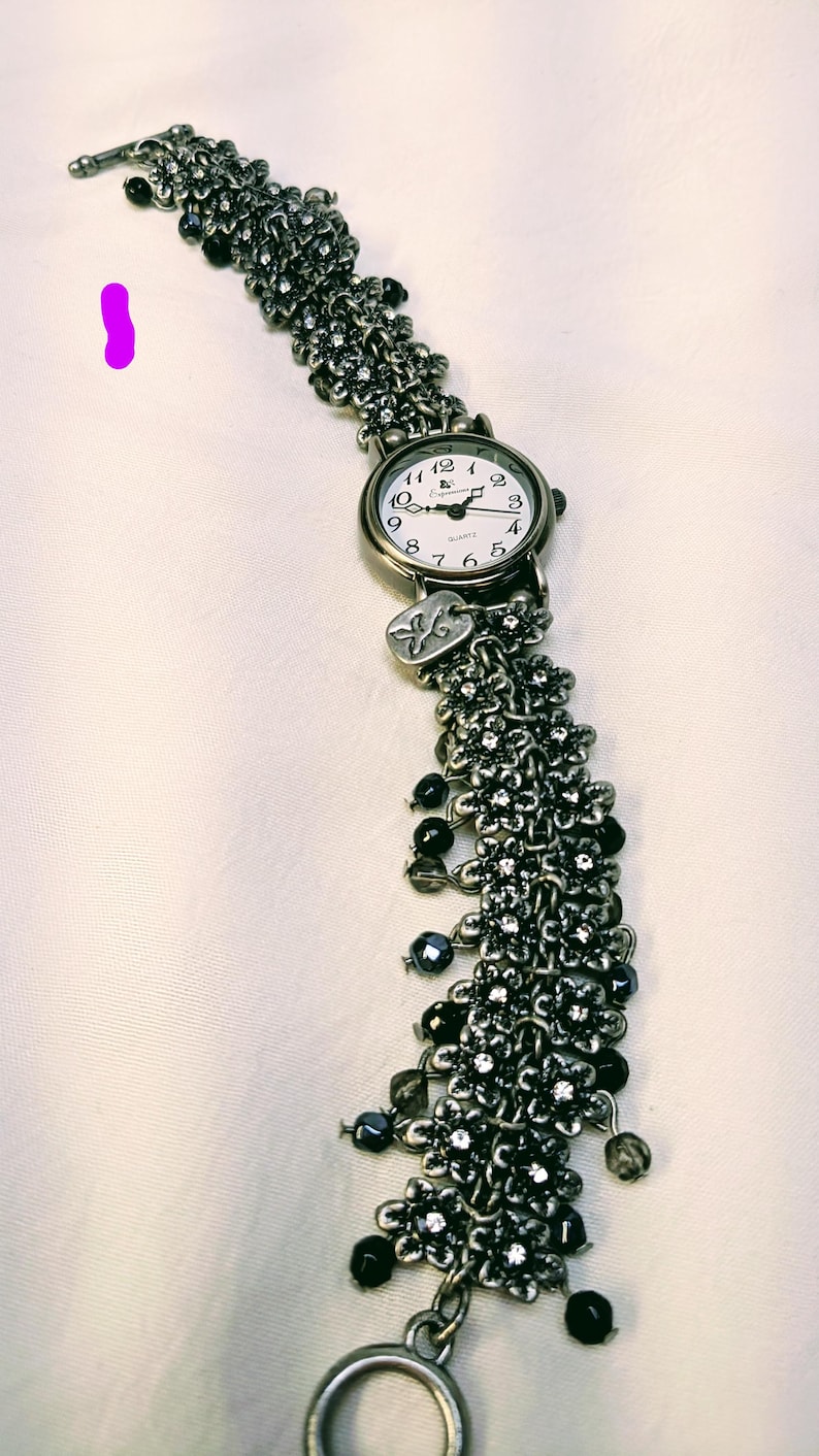 May include: A silver-toned quartz watch with a white face and black numerals. The band is composed of multiple chains adorned with small metal flower charms, clear crystals, and dangling black beads. A small rectangular metal tag hangs near the watch face.