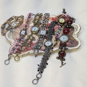 May include: A collection of six decorative wristwatches with varied bands, including beaded, metallic, and floral-accented designs, arranged on an ornate, rectangular, cream-colored tray featuring a central floral pattern with gold-tone trim.