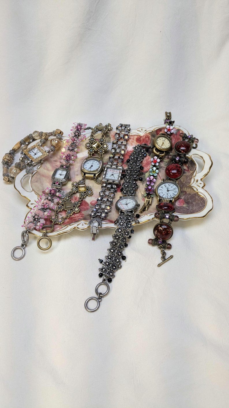 May include: A collection of six decorative wristwatches with varied bands, including beaded, metallic, and floral-accented designs, arranged on an ornate, rectangular, cream-colored tray featuring a central floral pattern with gold-tone trim.