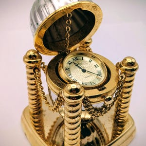May include: A gold-colored decorative clock with a silver top. The clock features a round face with Roman numerals and is suspended within a gold frame. Decorative chains and pillars add to the ornate design.