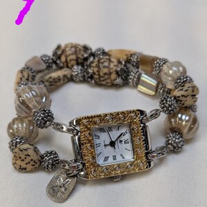 May include: A wrist watch with a square face featuring a gold-toned textured frame and Roman numerals. The watch band is composed of alternating tan speckled beads, clear ribbed glass beads, and small silver-toned metallic spacer beads.