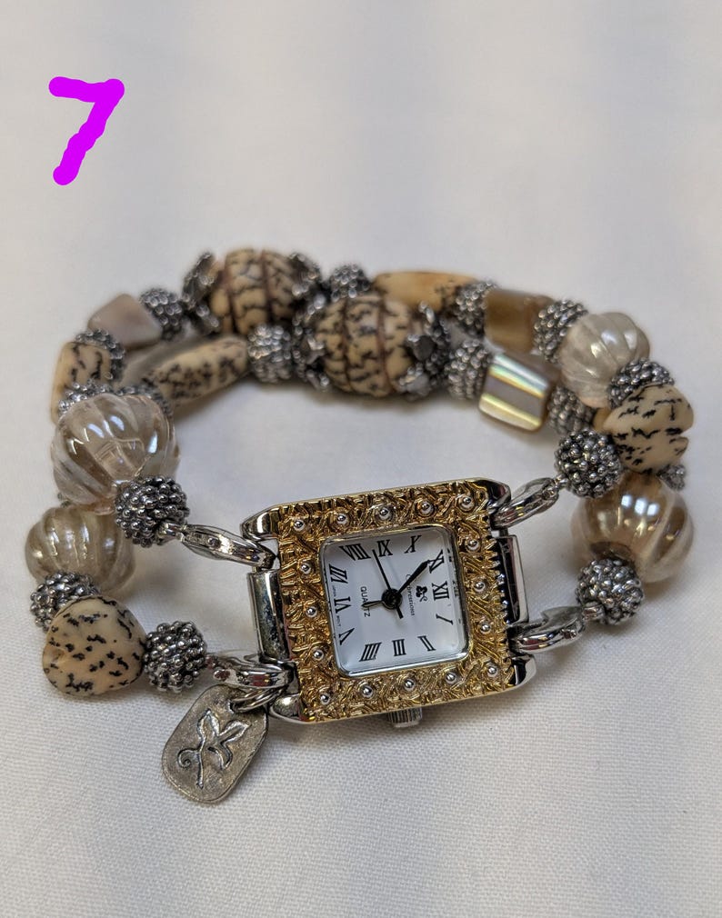 May include: A wrist watch with a square face featuring a gold-toned textured frame and Roman numerals. The watch band is composed of alternating tan speckled beads, clear ribbed glass beads, and small silver-toned metallic spacer beads.