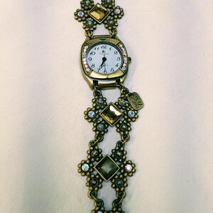 May include: A bronze-toned analog watch with a white face and black numerals. The band features ornate floral-shaped links, square amber-colored crystals, and small iridescent rhinestones. A small metal tag hangs from the watch casing.