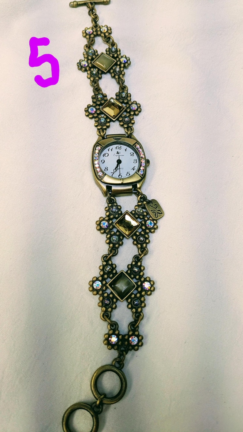 May include: A bronze-toned analog watch with a white face and black numerals. The band features ornate floral-shaped links, square amber-colored crystals, and small iridescent rhinestones. A small metal tag hangs from the watch casing.