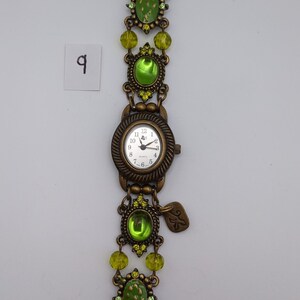 May include: A bronze-toned analog wristwatch featuring a round white face with black numerals and hands. The band is composed of ornate metal links, lime green oval cabochons, and faceted lime green beads. A small metal charm hangs near the watch face.