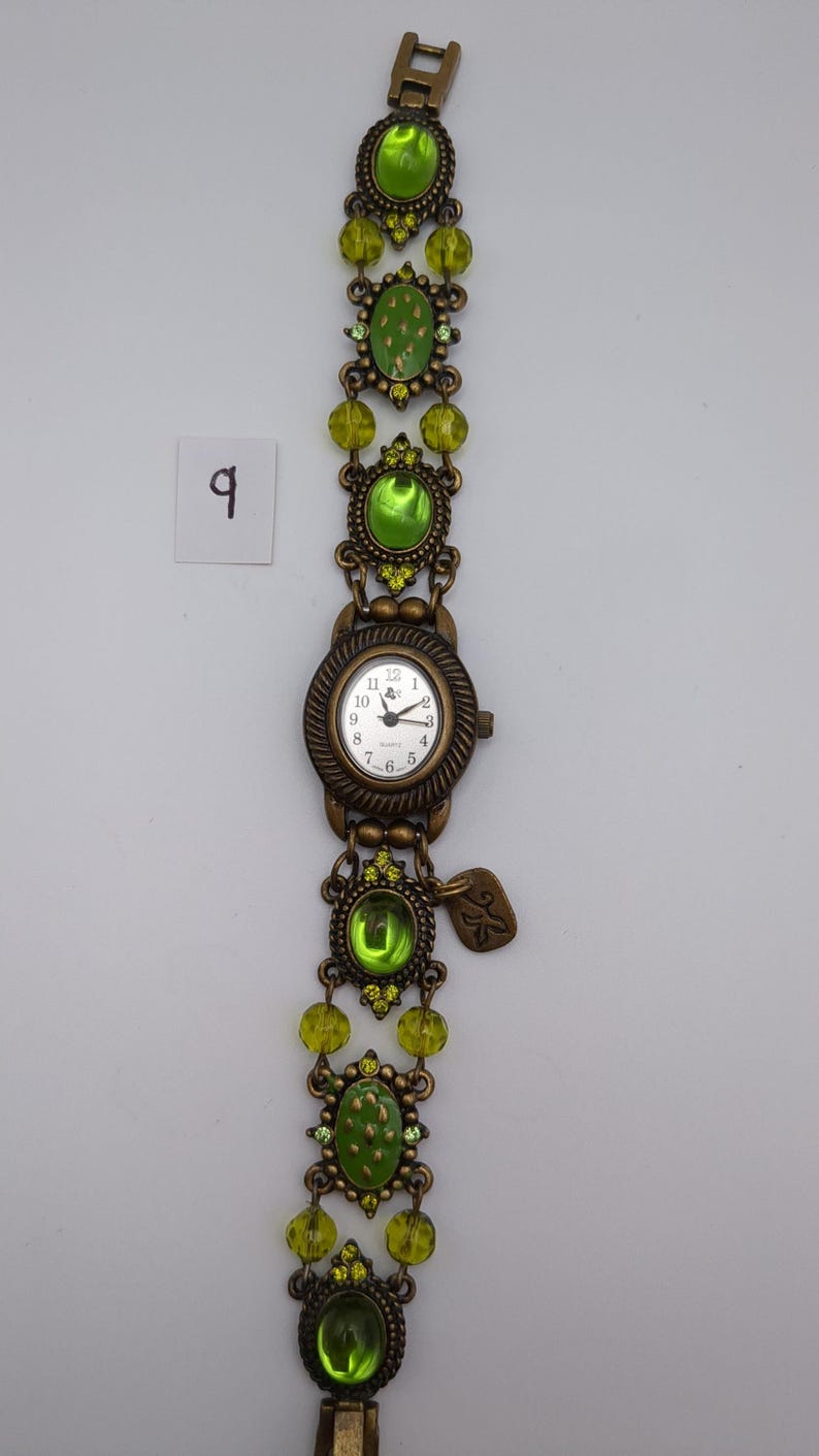 May include: A bronze-toned analog wristwatch featuring a round white face with black numerals and hands. The band is composed of ornate metal links, lime green oval cabochons, and faceted lime green beads. A small metal charm hangs near the watch face.