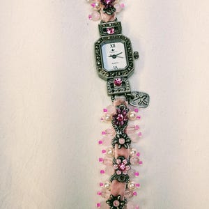 May include: A wrist watch with a rectangular white face, black hands, and Roman numerals. The band is made of pink velvet ribbon adorned with silver-toned metal floral accents, clear pink beads, and small white pearls, featuring a toggle clasp.