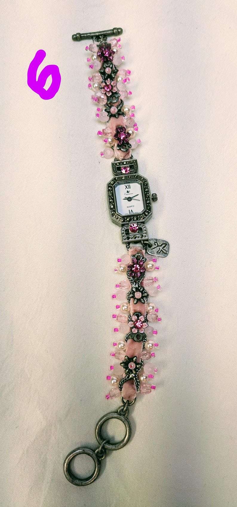 May include: A wrist watch with a rectangular white face, black hands, and Roman numerals. The band is made of pink velvet ribbon adorned with silver-toned metal floral accents, clear pink beads, and small white pearls, featuring a toggle clasp.