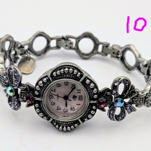 May include: A silver-toned quartz watch with a round face featuring a light pink dial and black numerals. The band consists of ornate metal links decorated with small purple crystals, iridescent stones, and dark red accents. The number 10 is visible.