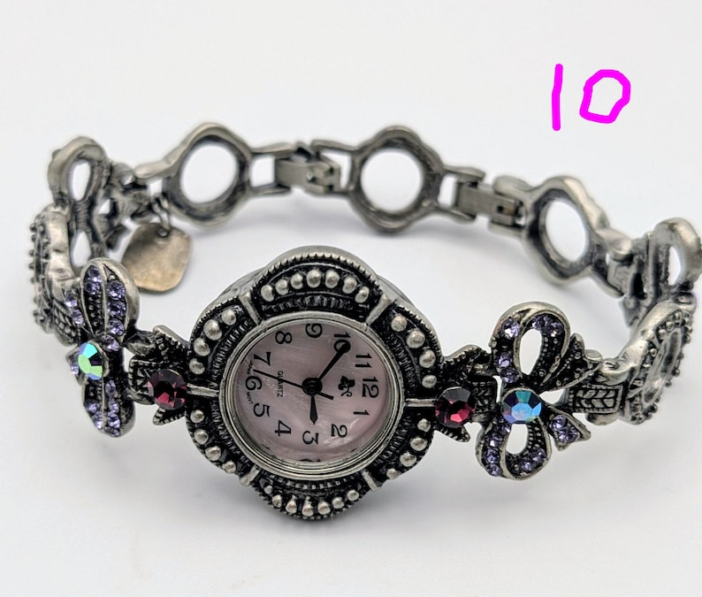 May include: A silver-toned quartz watch with a round face featuring a light pink dial and black numerals. The band consists of ornate metal links decorated with small purple crystals, iridescent stones, and dark red accents. The number 10 is visible.